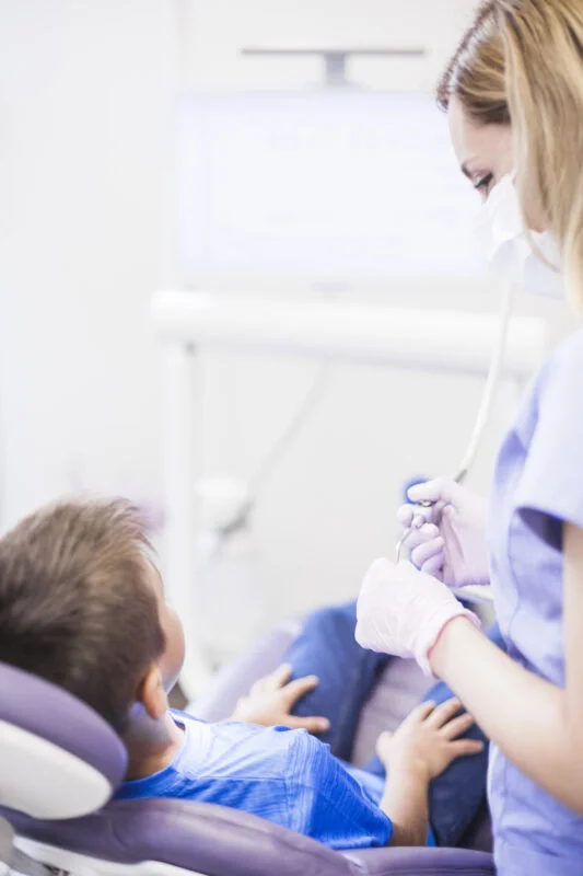 Woman With Ultrasonic Scaler Standing Near Boy Sitting Dental Chair